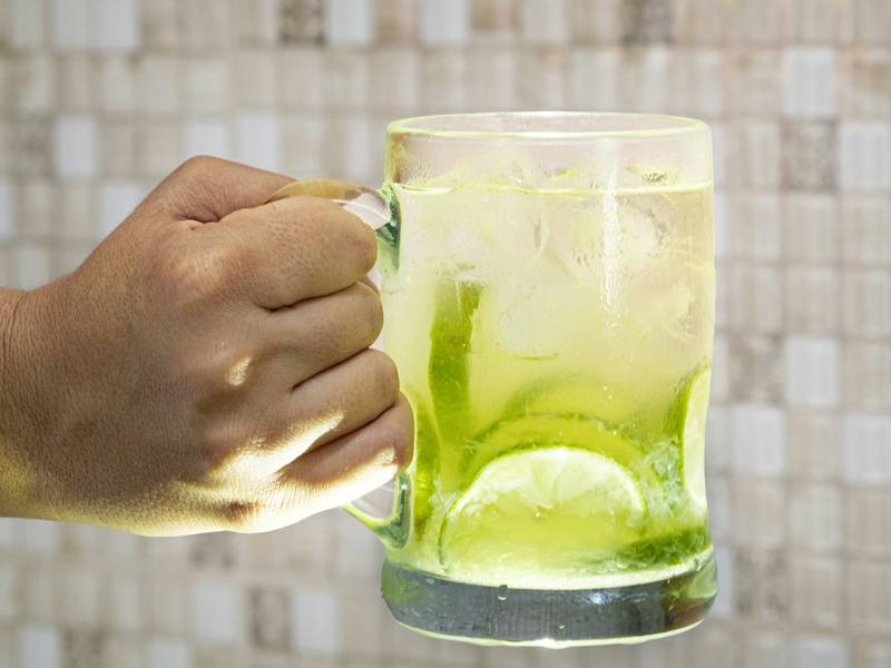 Mão segurando caneca de vidro com caipirinha de limão e gelo