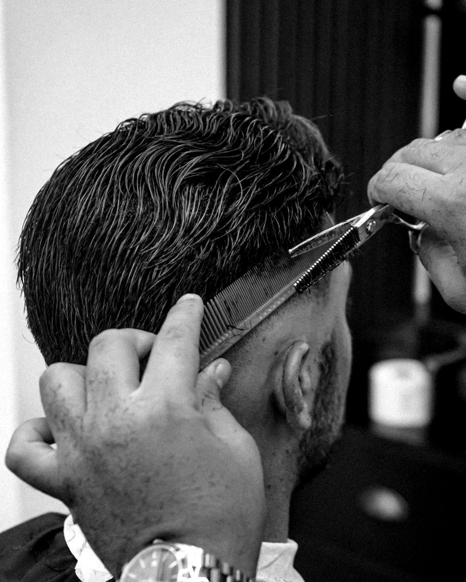 Barbeiro aparando a lateral do cabelo de um cliente com tesoura e pente, em preto e branco