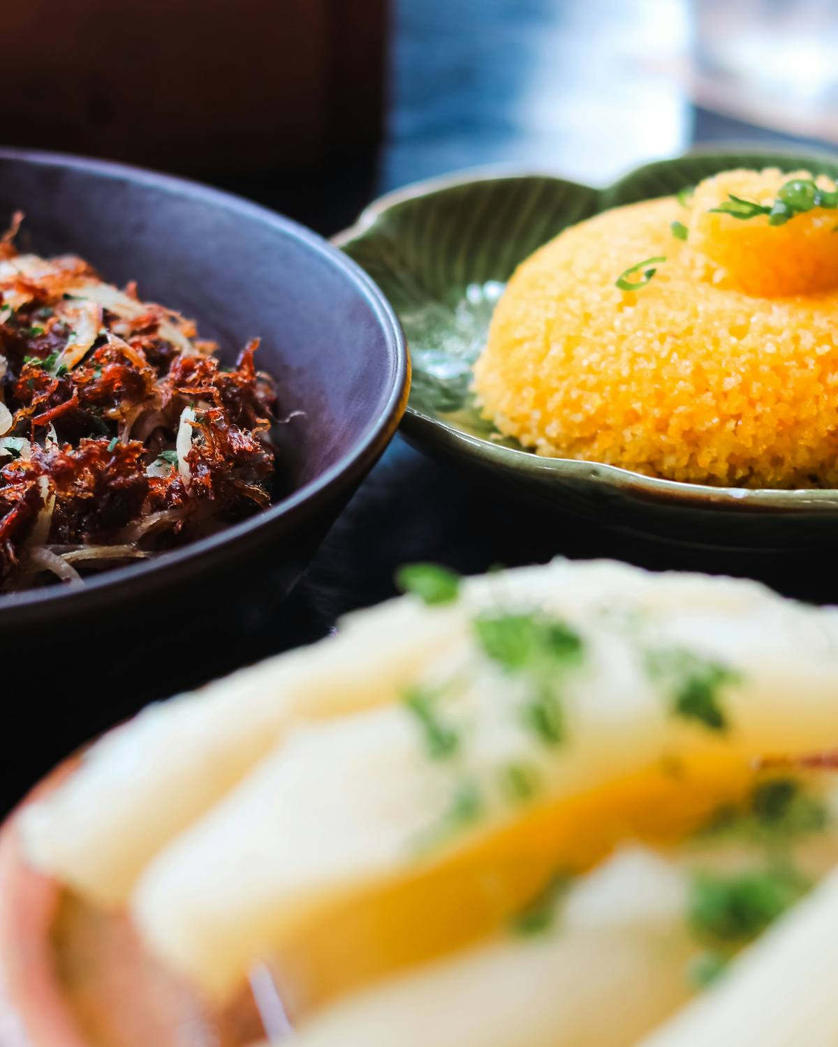 Mesa com carne-seca acebolada em tigela escura, cuscuz de milho em prato verde e aipim cozido em primeiro plano