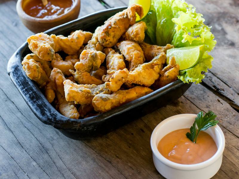 Iscas de peixe empanadas em travessa preta, com limão, alface e molho em potinho